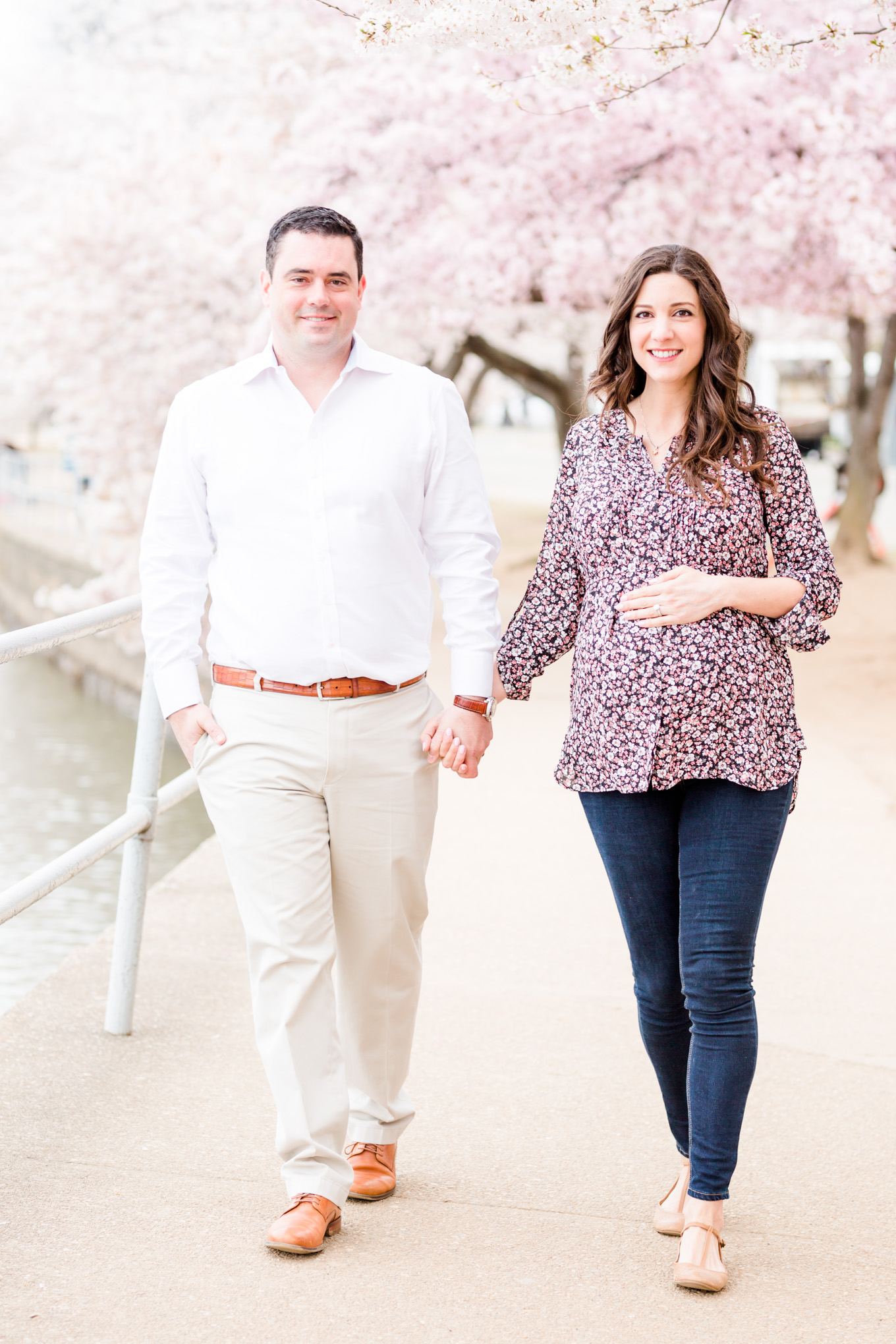 D.C. cherry blossoms maternity session, expectant couple, D.C. couple, maternity photos, maternity portraits, spring maternity photos, cherry blossoms, D.C. cherry blossoms, tidal basin, couple walking, couple holding hands, brunette mom to be, brunette woman, floral print top, dark wash blue jeans, 