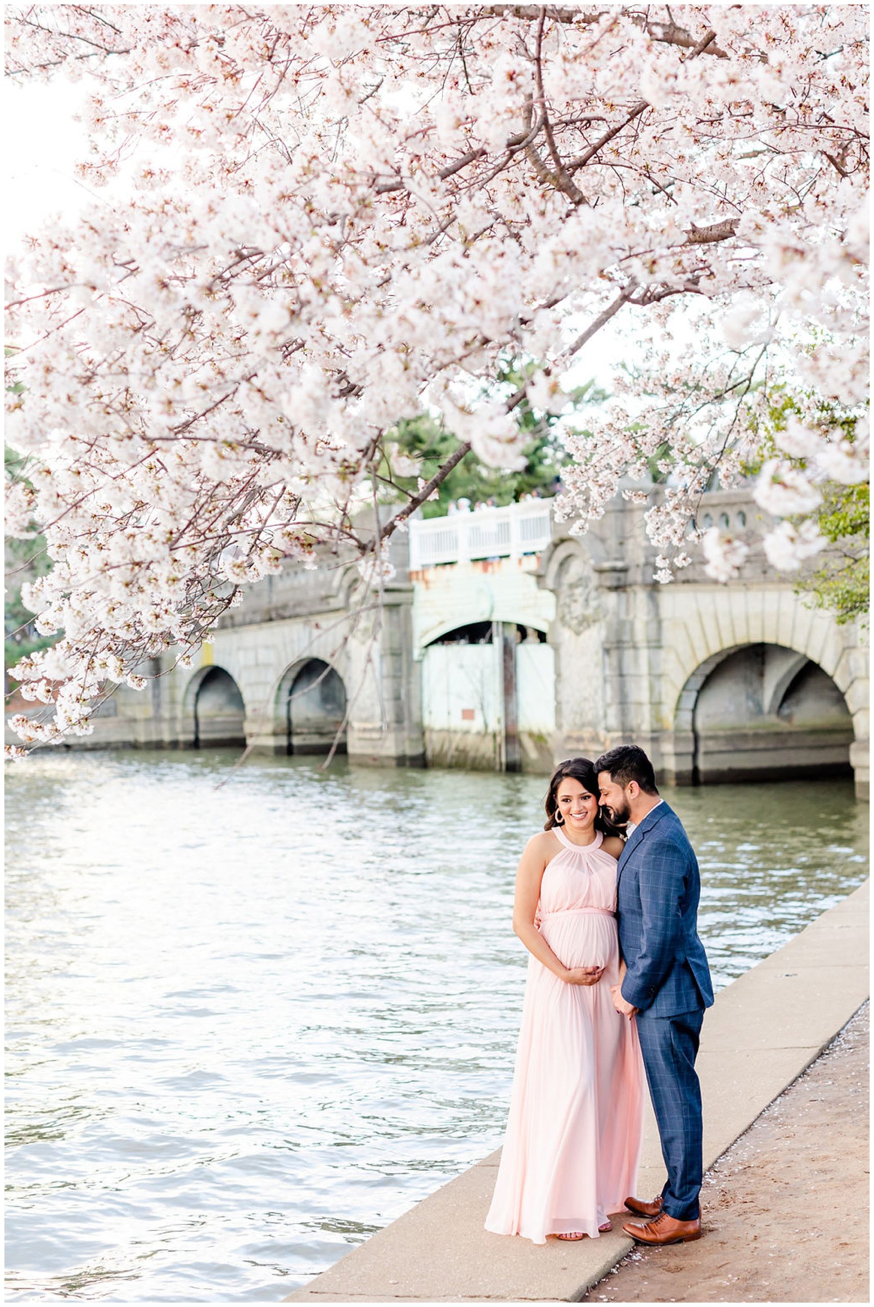 windy cherry blossoms maternity session, D.C. maternity photos, cherry blossoms maternity photos, D.C. portraits, D.C. cherry blossoms portraits, Tidal Basin Washington D.C., D.C. maternity photographer, D.C. photographer, spring portratis, Rachel E.H. Photography, couple in front of bridge, navy suit, woman holding baby bump